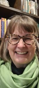 Sara Hood smiling at camera, earinag green scarf, with bookshelves behind